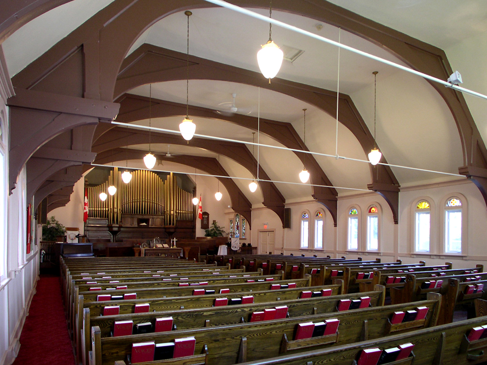 Photographie couleur, plan éloigné, intérieur d’une église dont les poutres de bois du bâtiment sont apparentes, en avant-plan, des bancs en bois contenant des livres rouges, en arrière-plan, un très grand orgue à tuyaux en cuivre. 