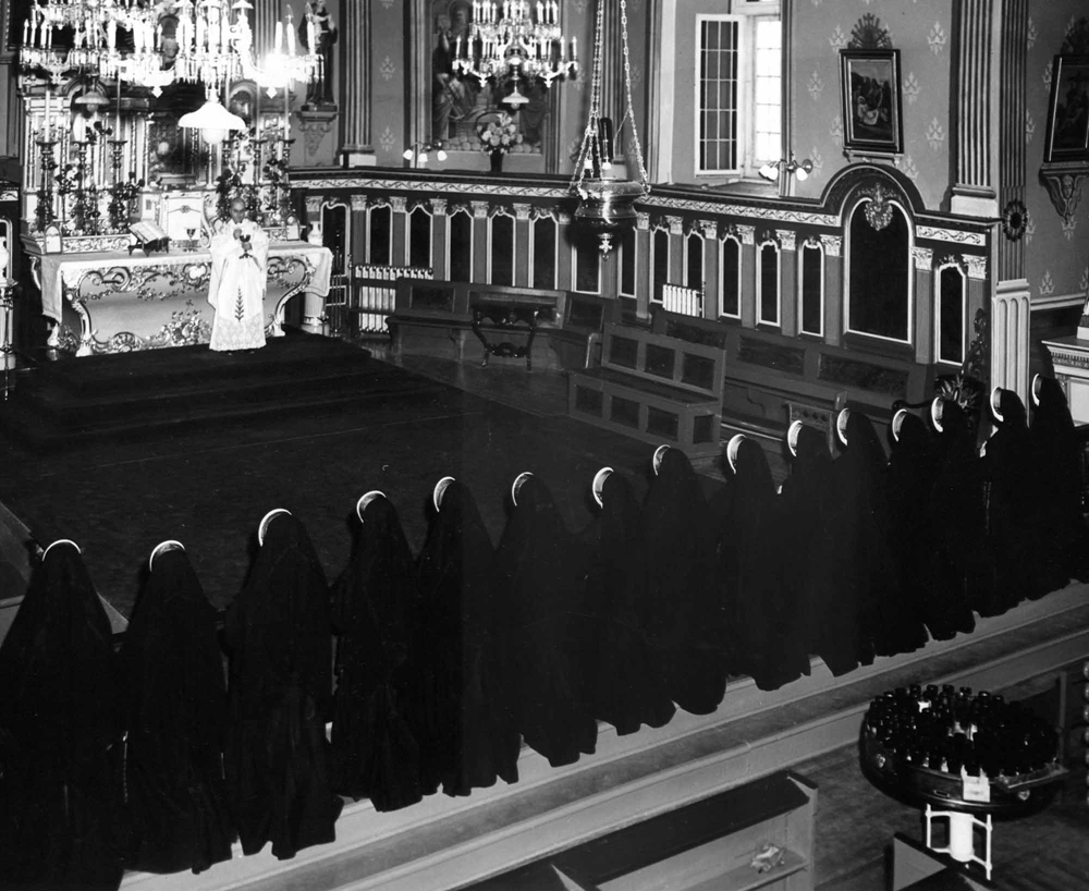 Photographie ancienne en noir et blanc, plan éloigné, intérieur d’une église richement décorée, en avant-plan, des religieuses vêtues de noir sont agenouillées devant un célébrant lors d’une cérémonie religieuse. 