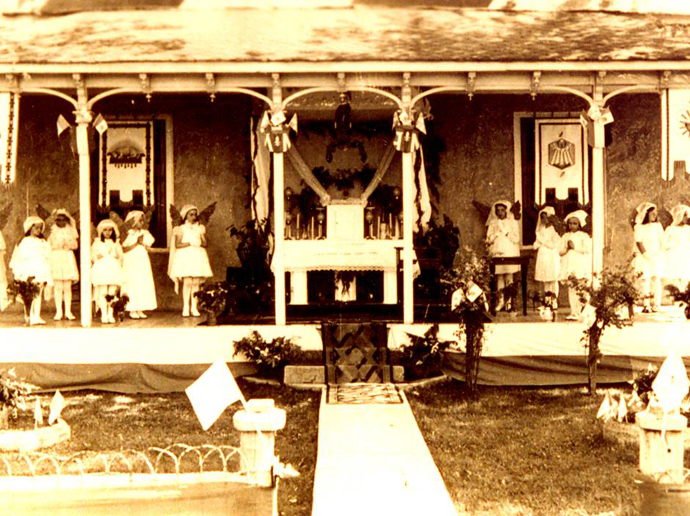 Photographie ancienne couleur sépia, devant une maison décorée de drapeaux et d’accessoires religieux, des fillettes habillées en blanc, les mains jointes, personnifient des anges de chaque côté d’un reposoir.