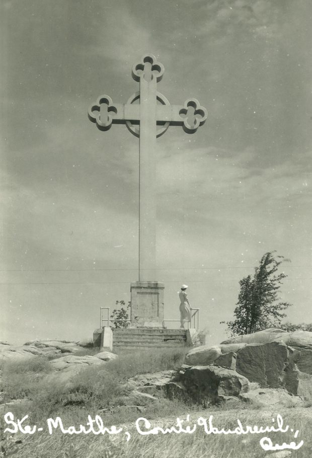 Photographie en noir et blanc, plan éloigné, par un jour de grand vent, une femme se tient debout et regarde au loin sous une immense croix de pierre installée sur un promontoire rocheux.