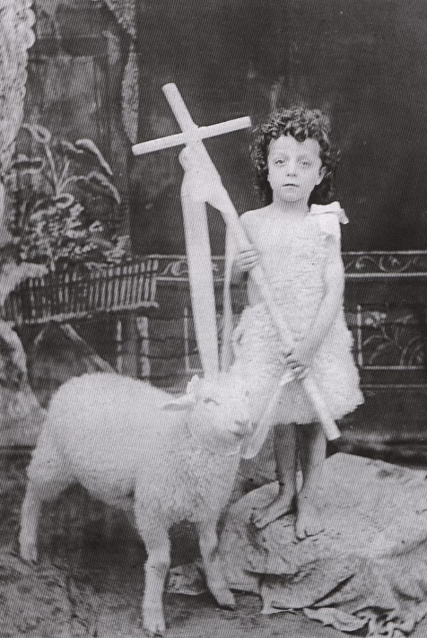 Photographie ancienne noir et blanc, plan rapproché, un petit garçon tenant une croix et vêtu d’une peau de mouton se tient debout à côté d’un petit mouton.