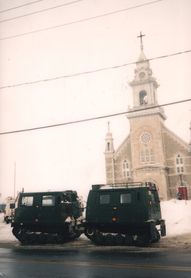 Photographie couleur prise en hiver, plan éloigné, deux chenillettes militaires sont stationnées devant la porte principale d’une grande église en pierres taillées.