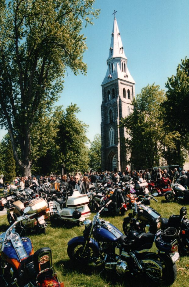 Photographie couleur, plan éloigné, des motos et une foule de personnes sont rassemblés sur un grand terrain entouré d’arbres devant une église. 