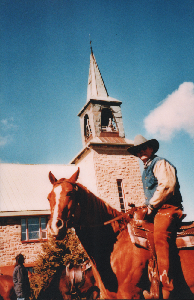 Photographie couleur, plan rapproché, un cowboy sur son cheval devant le clocher d’une église en pierres. 