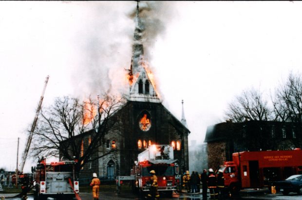 Photographie en couleur, plan éloigné, des pompiers, munis d’une grue et de plusieurs camions, tentent d’éteindre l’incendie d’une église dont le clocher, le toit et les fenêtres sont traversés par des flammes. 