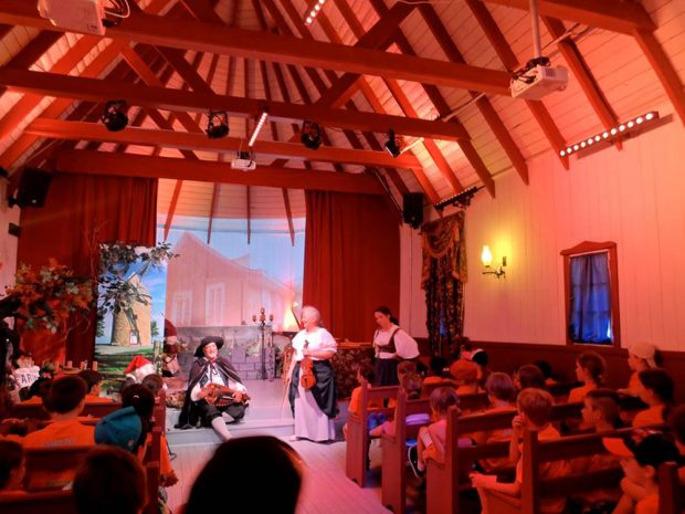 Photographie couleur, intérieur d’une petite chapelle convertie en salle de spectacle, des enfants assis sur des bancs en bois assistent à une représentation théâtrale avec des musiciens en costumes d’époque. 