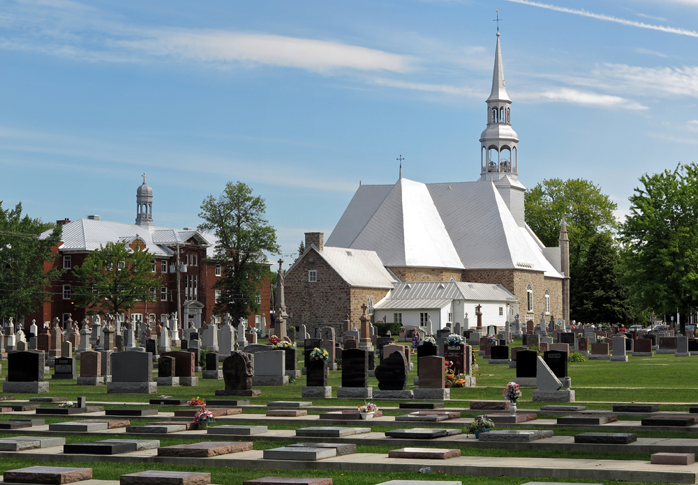 Photographie couleur, plan éloigné, en avant-plan, un grand cimetière, à l’arrière-plan, une église en pierres et un clocher, à la gauche du bâtiment, un grand édifice en briques rouges surmonté d’une coupole.