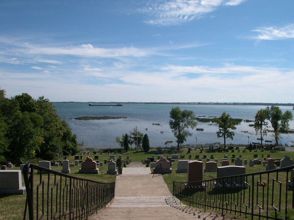 Photographie couleur, plan éloigné, à l’avant-plan, un long escalier descend au milieu d’un cimetière vers un vaste cours d’eau, en arrière-plan.