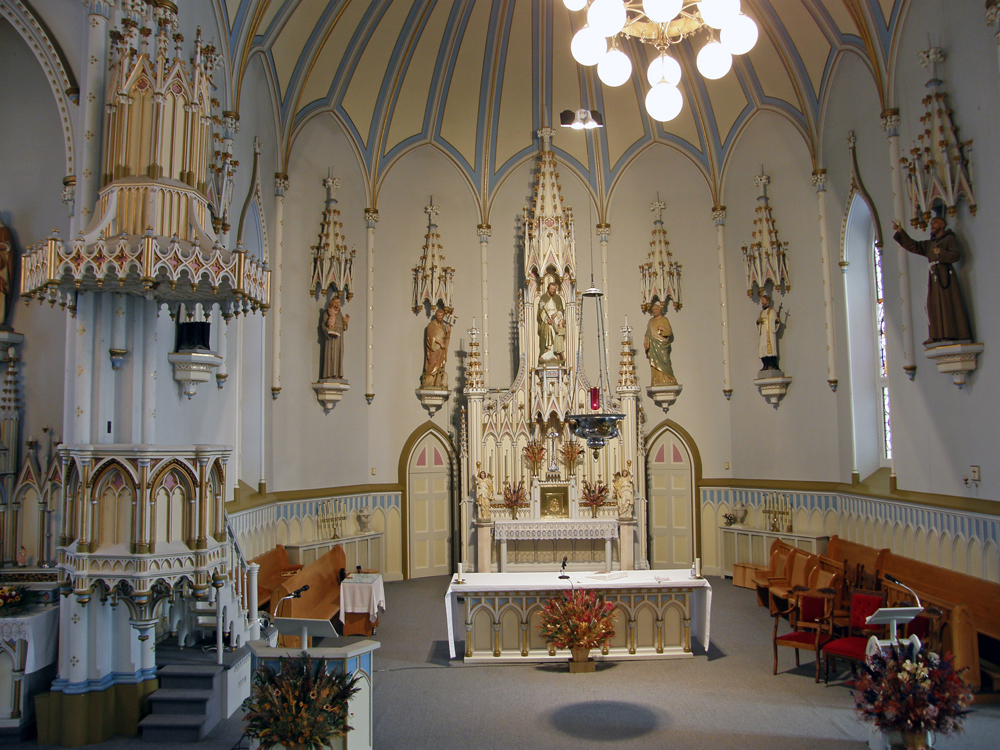 Vue intérieure d’une partie d’une église en demi-cercle,  plan éloigné, sur les murs plusieurs statues religieuses en plâtre posées sur des socles en bois  avec au-dessus d’elles des dais en bois sculptés, au centre, du mobilier liturgique. 