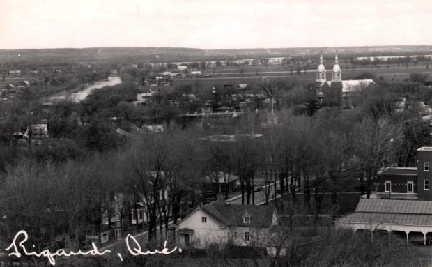 Photographie ancienne en noir et blanc, prise en hauteur, plan éloigné, vue sur les clochers d’une église qui percent entre les arbres et les maisons, en arrière-plan, une rivière.