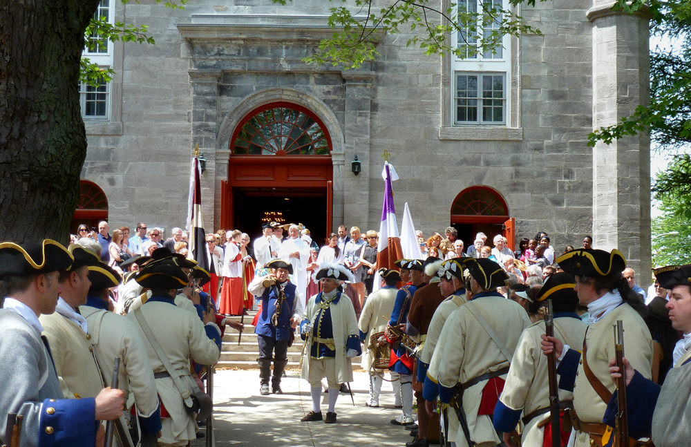 Photographie couleur, plan éloigné, une foule de personnes portant différents costumes d’époque sont sur le parvis d’une église en pierres à la sortie de la messe dominicale. 