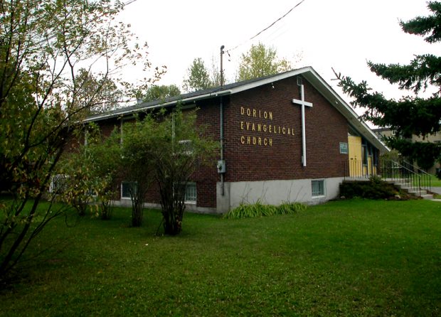 Photographie couleur, plan éloigné, vue latérale de la façade d’un bâtiment en briques brunes avec un toit à deux versants, sur la façade, une grande croix blanche à côté de laquelle on peut lire l’inscription Dorion Evangelical Church.