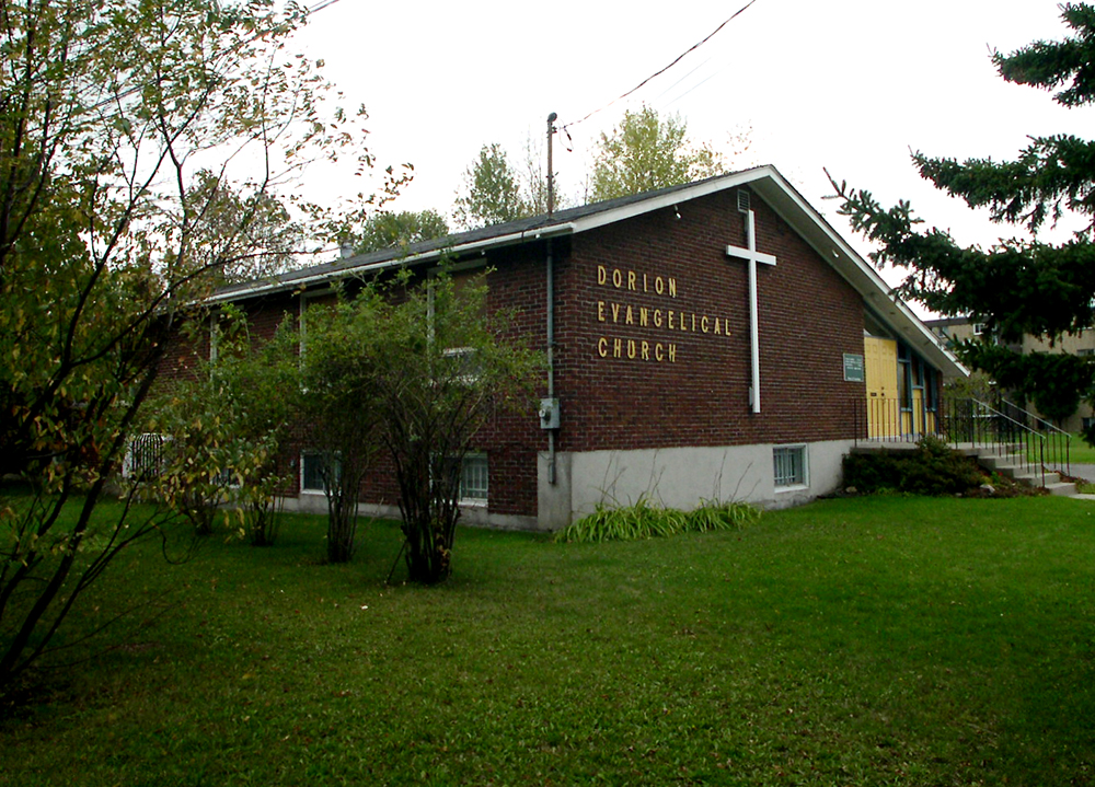 Photographie couleur, plan éloigné, vue latérale de la façade d’un bâtiment en briques brunes avec un toit à deux versants, sur la façade, une grande croix blanche à côté de laquelle on peut lire l’inscription Dorion Evangelical Church.