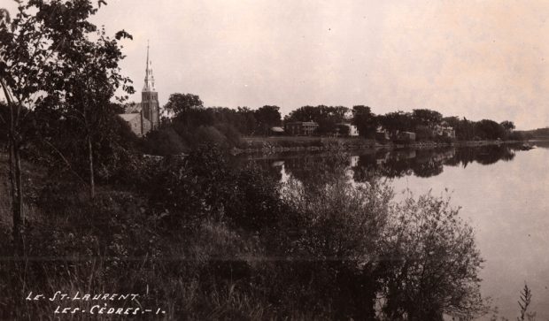 Photographie ancienne en noir et blanc, plan éloigné, en avant-plan, une vaste étendue d’eau, des arbres et de la végétation, en arrière-plan, le clocher d’une église avec à sa droite une série de petites maisons. 