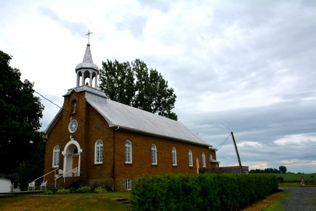 Photographie couleur, une église en briques rouges comprenant le clocher et une statue dans une niche, sur le parvis de l’église des chaises de jardin, sur le côté droit du bâtiment une fenêtre placardée et une annexe en plaques de béton, à l’arrière plan des champs.