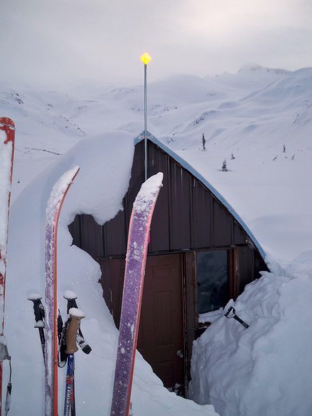 Des spatules et des bâtons de ski de couleur violette et rose coincés dans la neige à gauche de l’entrée et de la fenêtre avant partiellement déneigées du refuge voûté de style gothique.