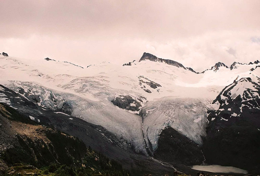 Un énorme glacier recouvre le versant et masque les sommets au-dessus. Le fluage du glacier s’étend jusqu’au fond de la vallée alpine en suspension. Des nuages gris tourbillonnent au-dessus des sommets élevés.