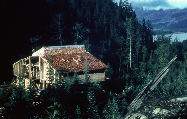 La finition de la charpente du grand chalet refuge est effectuée à l’aide de planchettes du côté droit du toit. Une feuille de plastique recouvre l’autre moitié du toit et la charpente dénudée.