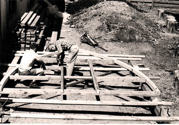 La charpente voûtée n’est plus à même le sol, mais soutenue par deux autres planches. Deux hommes courbés travaillant sur des éléments de la voûte sur le côté gauche. Une autre planche est soigneusement rangée derrière, à proximité d’une maison.