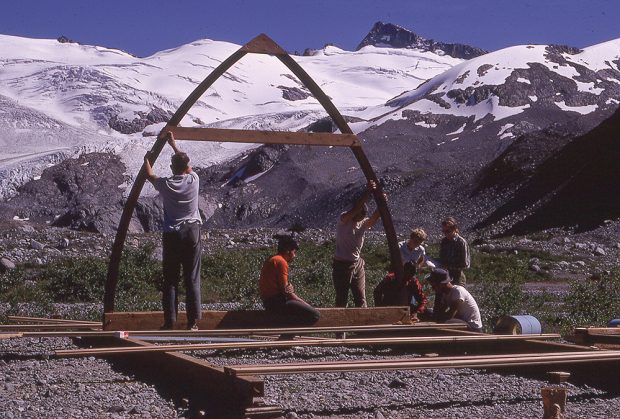 Les fondations sont achevées et plusieurs compartiments du plancher sont posés à l’horizontale au-dessus du sol rocheux. Deux hommes soutiennent la première voûte en bois, un autre est assis au milieu de celle-ci, tandis qu’un autre groupe s’emploie à fixer cette voûte à la fondation.