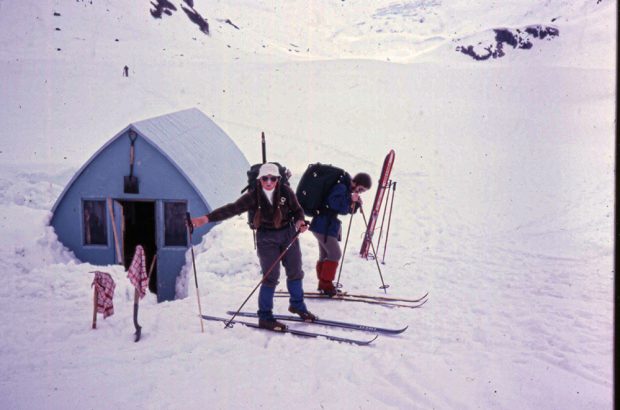 Une femme en chapeau blanc, chaussée de ses lunettes et de skis et tressée en longue natte se tient à côté d’un homme portant un sac à dos noir et une veste bleu, chaussant ses skis. Le refuge est visible derrière et l’entrée principale a été dégagée de toute couche de neige.