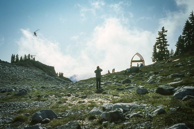 Un hélicoptère transportant une cargaison de fournitures survole l’extrême gauche du refuge; à l’avant, un autre membre prend une photo et à distance on aperçoit la structure voûtée et dénudée de la charpente, ainsi que les autres membres du club UBC-VOC.