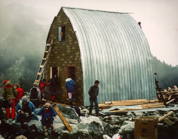 La structure du refuge est complétée à l’aide d’un revêtement en aluminium. Un homme regarde à travers la fenêtre de l’étage supérieur située au-dessus de l’entrée principale. D’autres membres travaillent sur le mur d’extrémité avant et devant l’entrée du refuge.