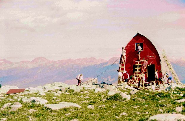 La structure du refuge a été construite et les membres du club UBC-VOC peignent la paroi d’extrémité avant en rouge vif. Les autres membres travaillent devant le refuge sous les rayons du soleil et des pics montagneux dénudés sont visibles au loin.