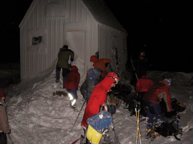 Trois membres déblayant la neige près de la porte principale. À leur extrême droite, un groupe de membres retirent leurs équipements de ski et rangent leurs effets.