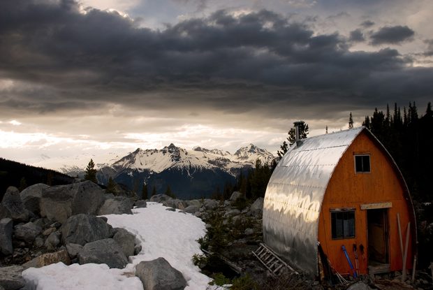 Le refuge déneigé muni d’un mur d’extrémité orange vif et d’un parement d’une couleur argent vif faisant miroiter les rayons du soleil. Le pré alpin visible derrière est parsemé de petits galets, d’arbres à feuilles persistantes et de plaques de neige. Au loin apparaissent des pics enneigés.