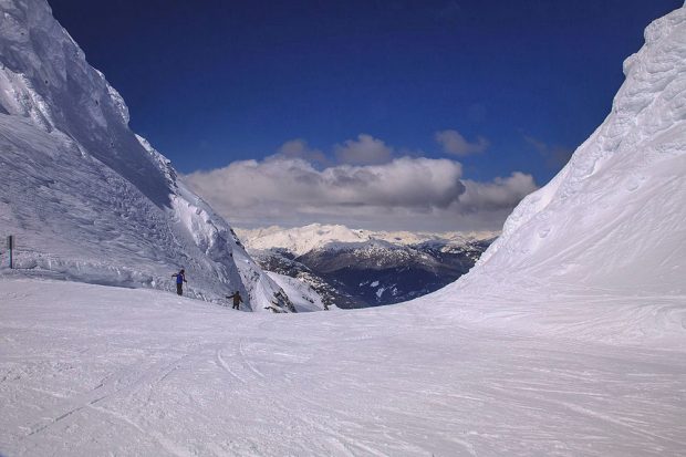 Deux grands murs de neige bordent une descente à ski escarpée et la neige réfléchit les rayons du soleil sous un ciel d’un bleu intense. On voit deux skieurs près de l’un des murs de neige à l’extrême gauche de la descente à ski.
