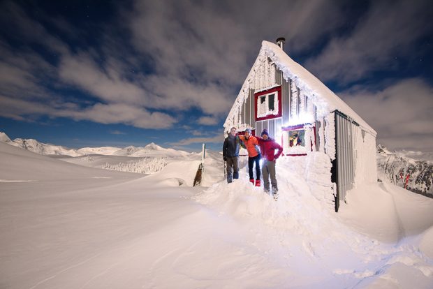 Un homme vêtu d’une veste bleu foncé et d’un pantalon gris, une femme portant une veste d’un orange éclatant et un pantalon noir et un homme portant un bonnet, une veste rouge et un pantalon gris clair posent devant le refuge bâti sur le mont Brew.