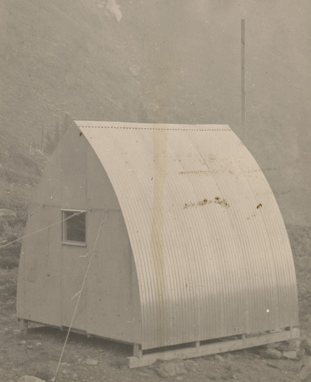 Vue arrière du parement métallique ondulé, refuge à voûte de style gothique comprenant une petite fenêtre carrée installée à l’emplacement prévu. En arrière-plan, un gros poteau et un escarpement rocailleux.