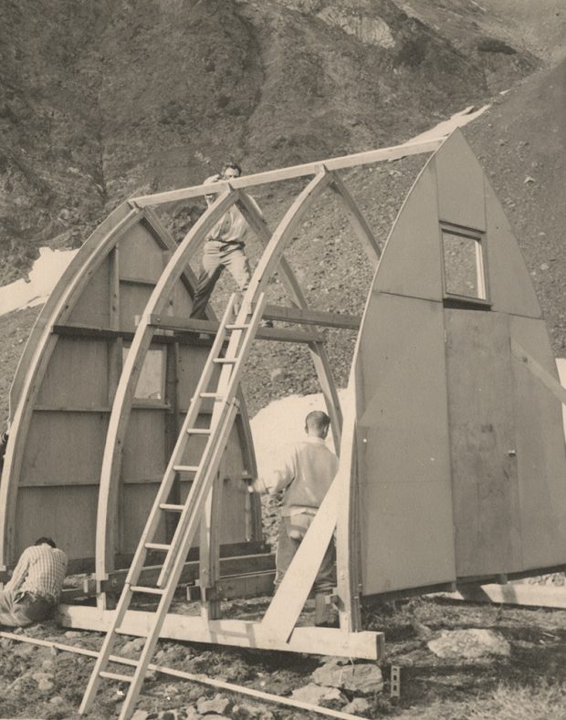 Trois hommes travaillent sur différents aspects de la charpente en bois voûtée du refuge de sorte à pouvoir installer le matériau de couverture.
