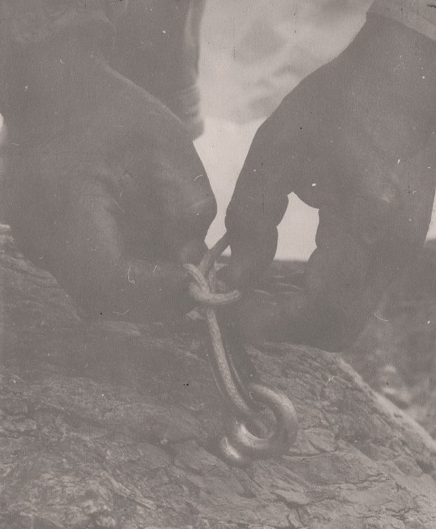 Gros plan sur des mains faisant des nœuds. Le câble décrit une boucle à travers un anneau incrusté dans le soubassement situé près du refuge.