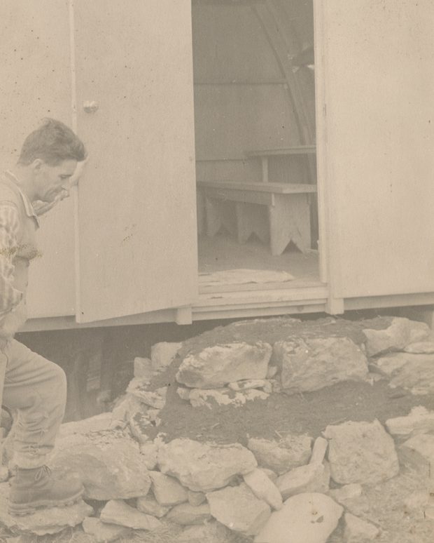 Un homme debout à gauche maintient la porte du refuge ouverte et inspecte les travaux qu'il a effectués à ladite entrée.