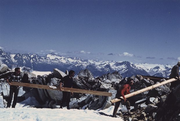 Deux membres de l’équipe transportent un long montant de deux par quatre tandis qu’un autre tient une longue planche. Des amas de neige couvrent le sol et des sommets enneigés sont visibles au loin.