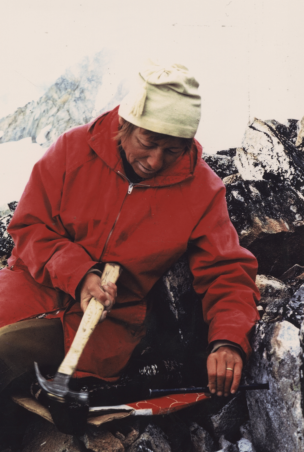 Une femme vêtue d’un manteau rouge et d’un bonnet blanc est assise, appuyée contre le rocher, et manie un marteau tout en tenant une longue pointe en métal incrustée dans le soubassement.