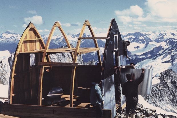 Le refuge Plummer est en cours de construction. Des hommes revêtent un mur d’aluminium. Un vaste paysage de montagnes enneigées est visible en arrière-plan.