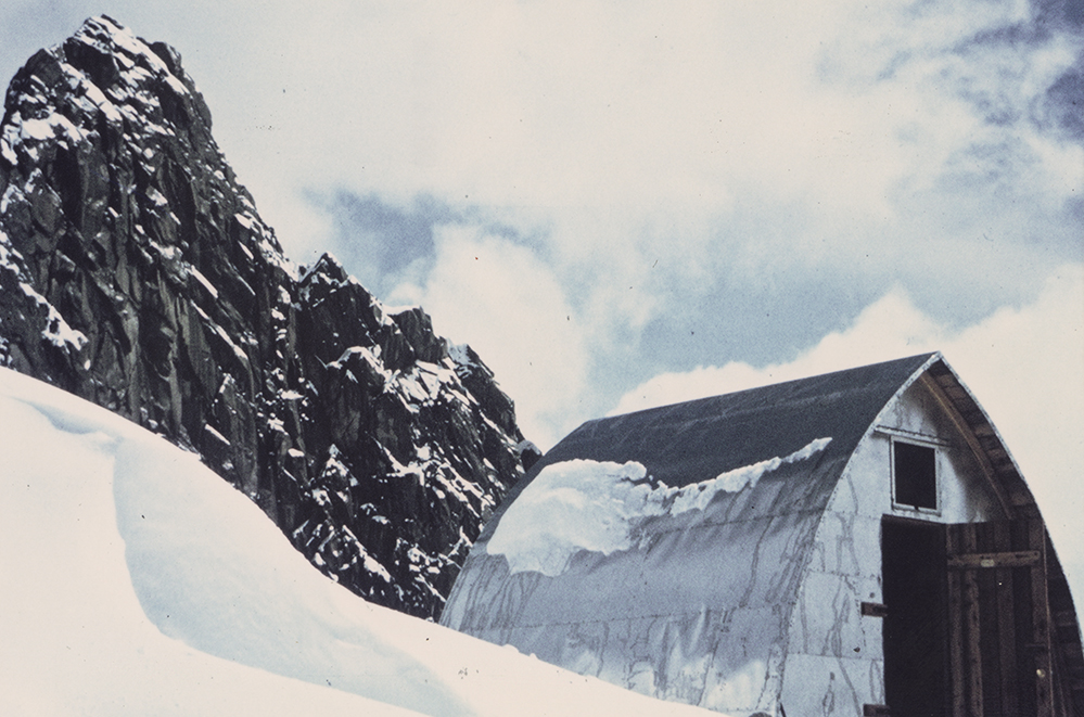 Un pic rocheux couvert d'une mince couche de neige est visible en arrière-plan et le refuge désormais achevé et entouré d'une neige épaisse se dresse au premier plan.