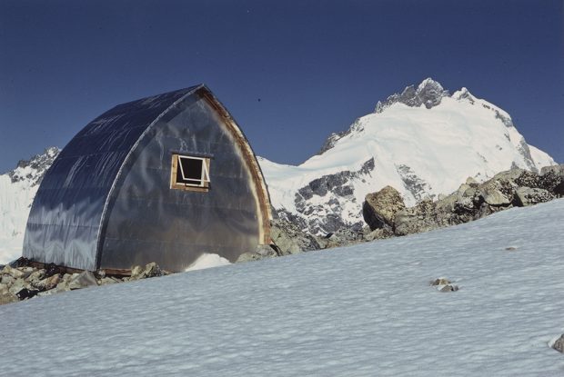 Une fine couche de neige couvre le versant arrière du refuge Plummer désormais achevé. Les alentours du refuge ont été complètement déblayés. En arrière-plan, un grand pic enneigé.