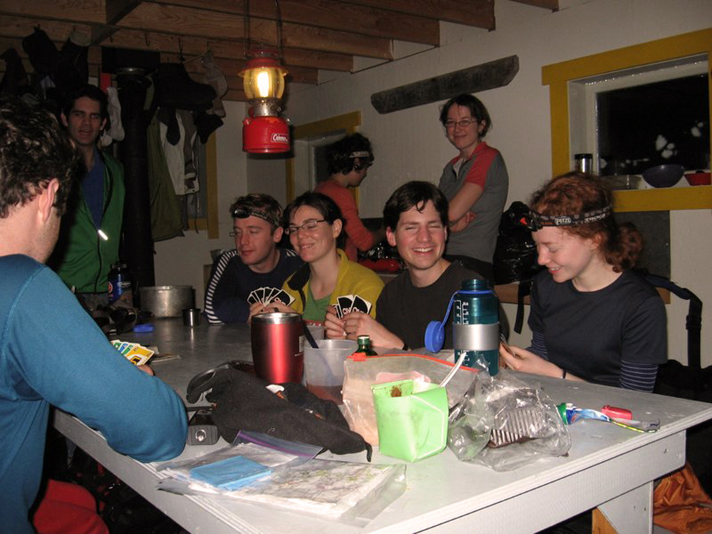 Rassemblés autour de la table à manger grise à l’intérieur du refuge, les membres du club jouent une partie de cartes UNO. Les autres membres debout en arrière-plan regardent la partie.