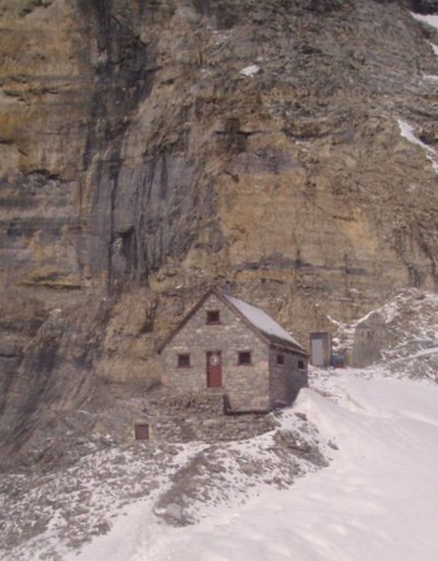 Un abri de pierres muni d’un toit en A et d’une porte centrale brun foncé se trouve en contrebas d’un rocher sédimentaire abrupt au milieu des montagnes Rocheuses. En arrière-plan, l’entrée des toilettes extérieures est creusée dans un rocher escarpé et à droite du refuge se trouve un versant enneigé qui longe jusqu’au bord de l’image.