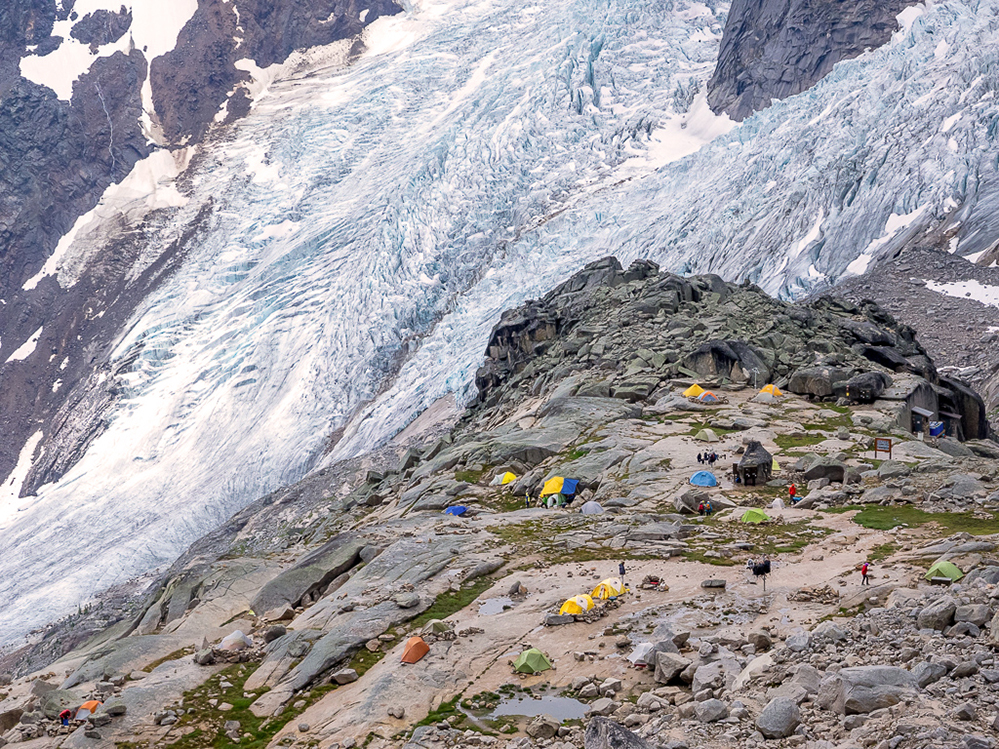 La photo offre une vue plongeante sur la grande zone rocheuse ouverte parsemée de tentes, à proximité d’un grand glacier contigu à une pente abrupte visible en arrière-plan.