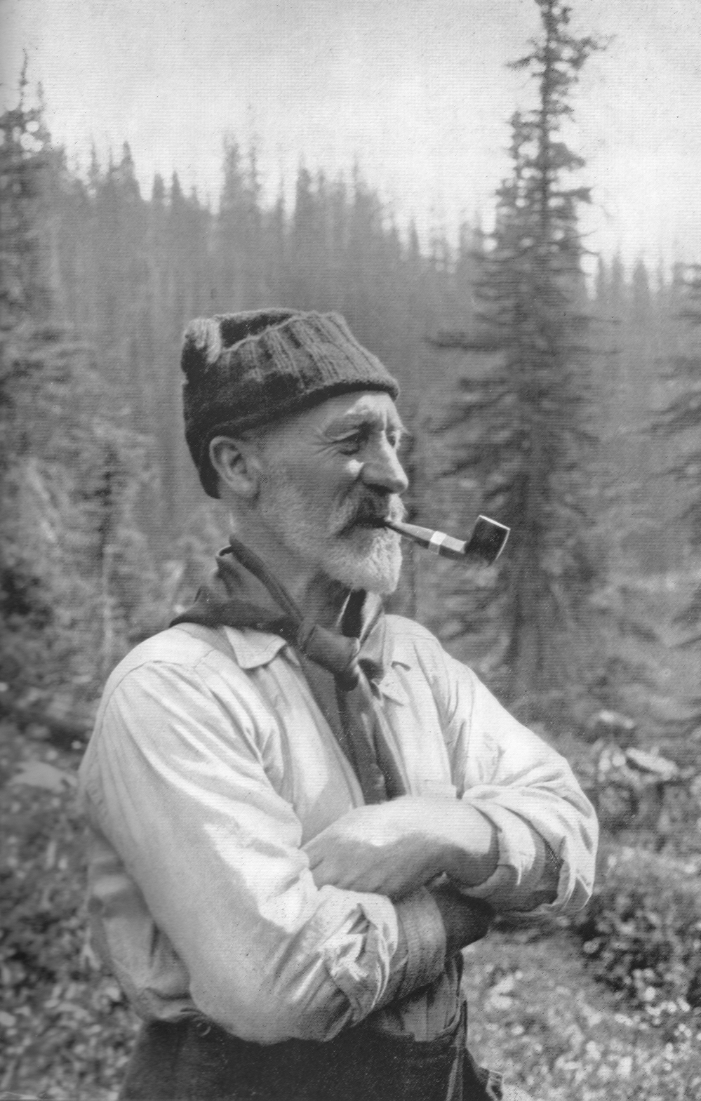 Une image en noir sur blanc d’Arthur Wheeler barbu, les bras croisés autour de la poitrine et une pipe dans la bouche, portant un bonnet, une écharpe et une chemise longue manche, et des arbres à feuilles persistantes en arrière-plan.