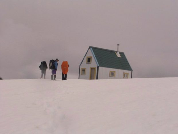 Trois membres portant de grands sacs à dos arpentent un versant enneigé se trouvant devant le refuge.  Des nuages gris planent sur le toit vert du refuge. 