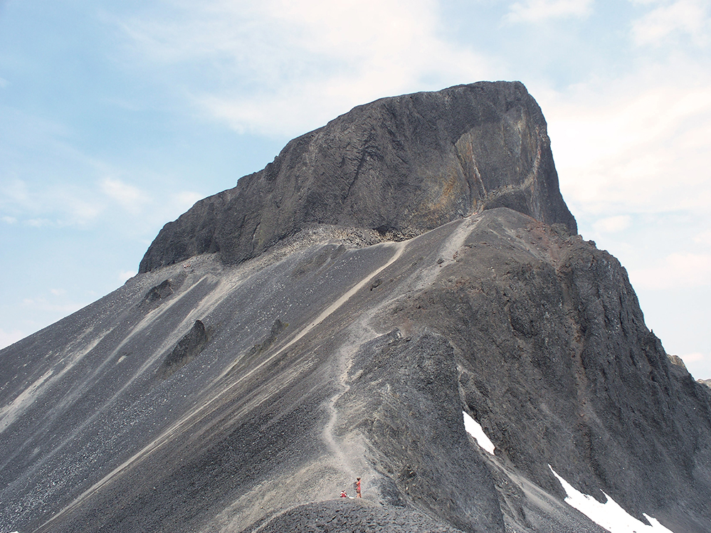 Gros plan du sombre rocher volcanique qui permet de reconnaître très facilement la crête de l’Aiguille. Une personne en chemise orange se tient près d’une crête sous l’aiguille.