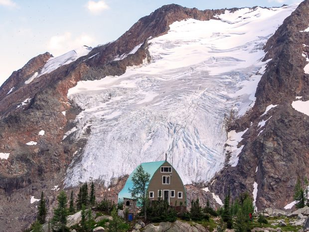 Le refuge au toit à voûte vert menthe dotée de nombreuses fenêtres et d’une porte à bordure blanche est entouré d’arbres à feuilles persistantes. Un grand glacier descend en cascade le long d’une pente abrupte derrière le refuge.