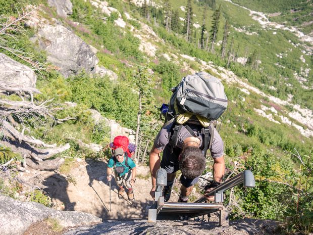 Photo prise de dessus avec vue plongeante sur un homme portant un sac à dos gris qui grimpe l’échelle. Un autre homme coiffé d'une casquette de baseball et portant un sac à dos rouge regarde d’en bas tout en tenant ses bâtons de randonnée.