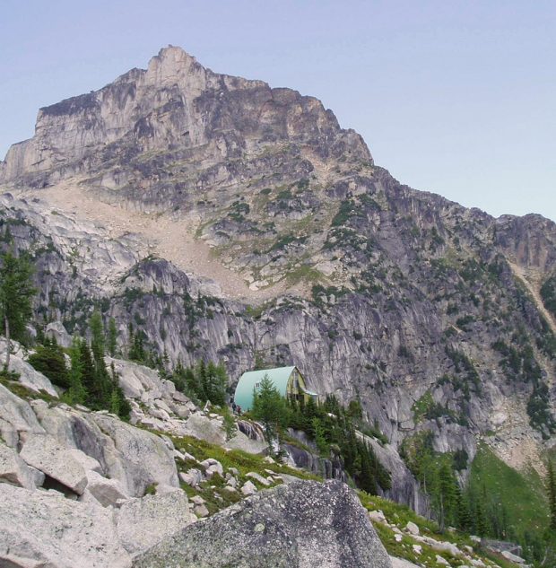 De bas en haut, le refuge Conrad Kain est entouré d’arbres à feuilles persistantes et la flèche de la grande arête des Bugaboos est visible à droite de la cabane.